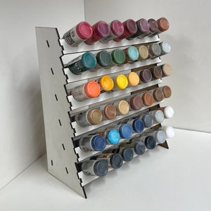 May include: A white wooden paint rack with multiple shelves holding rows of small paint bottles in various colours. The bottles are arranged in a staggered pattern, creating a visually appealing display.