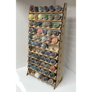 May include: A wooden paint rack with multiple shelves, holding rows of small paint bottles with various colours. The rack is designed to store and display paint bottles.