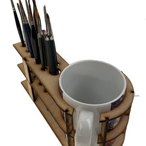 May include: Wooden paintbrush holder with a built-in mug holder. The holder has multiple slots for brushes and a space for a mug. The holder is made of light brown wood.