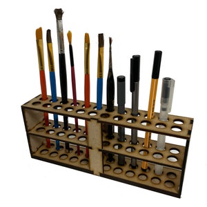 May include: A wooden art supply organiser with multiple holes to hold paint brushes, pens, and other art tools. The organiser is made of light brown wood and has a tiered design. Various paint brushes with red, blue, and yellow handles are visible.