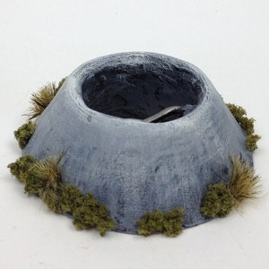 Op de afbeelding: Een klein, grijs, kraterachtig model met een donkergrijs interieur. Het model is bedekt met groene en bruine plukjes kunstgras.