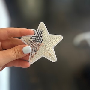 May include: A silver star-shaped patch with sequins. The patch is made of white felt and has a silver glitter finish.