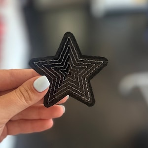 May include: A black star-shaped patch made of sequins. The star has multiple layers of sequins, creating a dimensional effect.