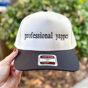 May include: A white and black baseball cap with the text "professional yapper" embroidered on the front. The cap has a black brim and a black button on top. The cap is held by a hand.