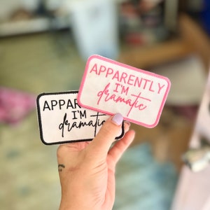 May include: Two white fabric patches with black and pink embroidered text. The top patch says "Apparently I'm dramatic" in pink lettering. The bottom patch says "Apparently I'm dramatic" in black lettering.