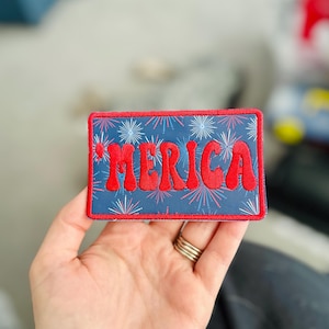 May include: Red embroidered patch with the word "'MERICA" in a retro font on a blue background with white and red firework designs.