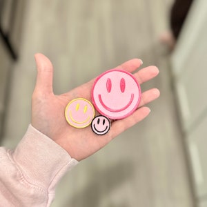 May include: Three embroidered smiley face patches. One is pink with a yellow outline, one is yellow with a pink outline, and one is black with a white outline.