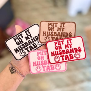 May include: Four embroidered iron-on patches with the text "Put it on my husband's tab" in different colors and fonts. The patches are on a hand.