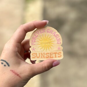 May include: A beige round sticker with a pink sun and the text "FOREVER CHASING SUNSETS" in pink lettering.