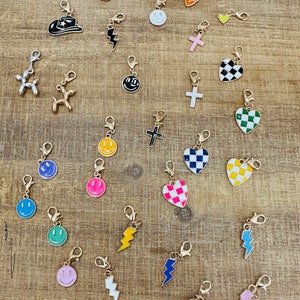 May include: A collection of colorful charms with gold-tone clasps. Charms include smiley faces, hearts, lightning bolts, a cowboy boot, a cowboy hat, a horseshoe, and a cross. The charms are in various colors, including pink, blue, yellow, and green.