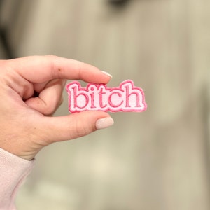 May include: Pink and white embroidered patch with the word "bitch" in white letters with a pink outline.