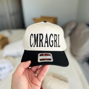 May include: A white and black baseball cap with the text "CMRAGRL" in black block letters. The cap has a black brim and a white crown. The cap is being held up by a hand.