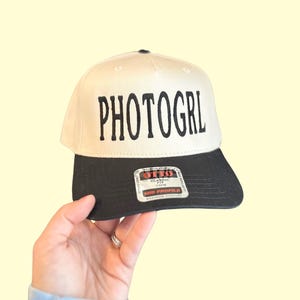 May include: A cream-colored baseball cap with a black brim and the word "PHOTOGRL" embroidered in black. The hat is held up by a hand against a light yellow background. The hat has a "Mid Profile" tag.