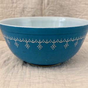 May include: A round mixing bowl with a matte teal exterior and a glossy white interior. The exterior features a white printed pattern consisting of a scalloped line along the upper rim with small starburst or snowflake motifs below the arches.
