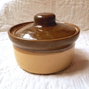 May include: A round ceramic casserole dish with a glossy dark brown lid and a smooth light beige base. The pot features a wide rim and a central knob on the lid, showcasing a two-tone glazed finish that covers the entire exterior surface.