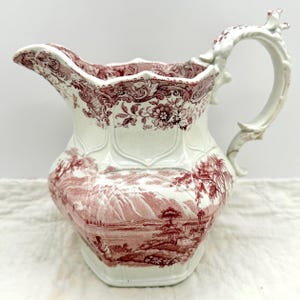 May include: A cream-colored ceramic pitcher featuring a deep red transferware pattern. The design includes a detailed landscape scene with mountains, trees, and buildings, complemented by an ornate floral border and a sculpted handle.