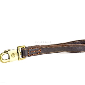 Puede incluir: Una correa de perro de cuero marrón con un cierre de latón y un diseño de huella de pata. La correa tiene costuras de color naranja.