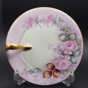 May include: A round, hand-painted porcelain plate with a gold handle. The plate features a pink and cream background with pink rose floral designs and gold trim. The plate is displayed on a clear stand.