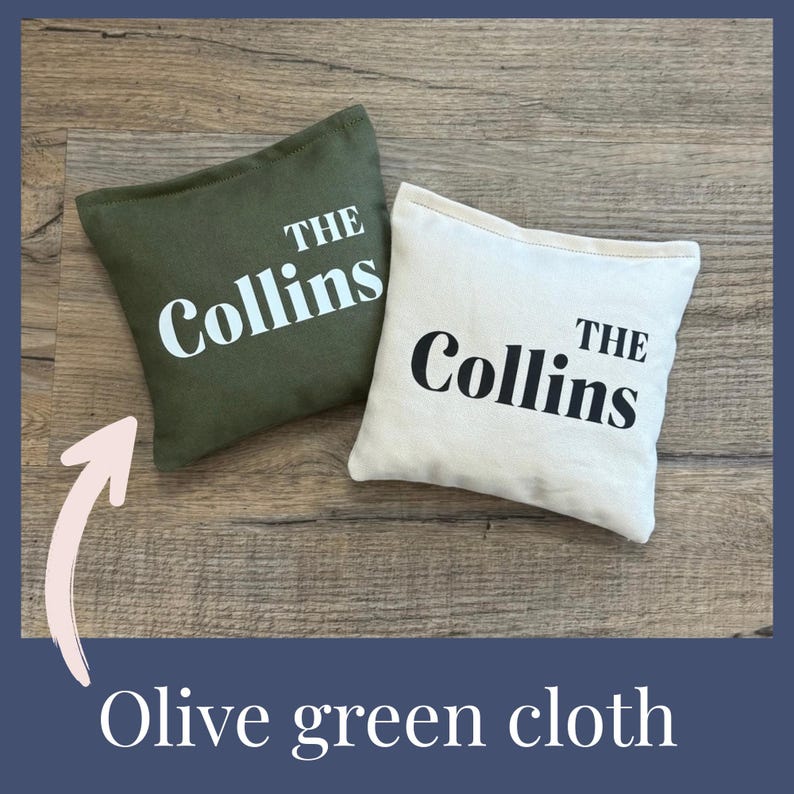 May include: Two small, square cloth pillows. One is olive green, the other is off-white. Both have the name "The Collins" printed in white and black, respectively. The pillows are on a wooden surface.