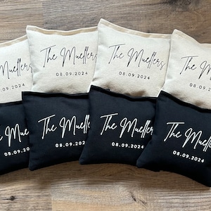 May include: A collection of personalized pillows, some in natural linen and others in black fabric. Each pillow features the text "The Muellers" and the date "08.09.2024" in elegant white script, ideal for wedding favors or home decor.