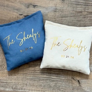 Puede incluir: Dos cojines personalizados, uno azul y otro blanco, con texto dorado "The Shealys" y una fecha. El coj&iacute;n azul tiene la fecha 07.21.26, y el blanco 02.21.26.