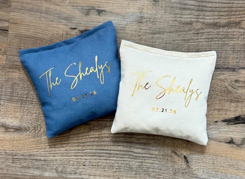 Puede incluir: Dos cojines personalizados, uno azul y otro blanco, con texto dorado "The Shealys" y una fecha. El coj&iacute;n azul tiene la fecha 07.21.26, y el blanco 02.21.26.