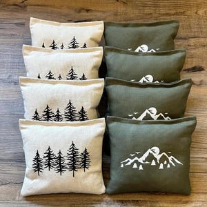 May include: Eight decorative throw pillows. Four are off-white with black tree designs, and four are olive green with white mountain and sun graphics. The pillows are rectangular and arranged on a wooden surface.