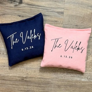Puede incluir: Dos cojines decorativos cuadrados. Uno es azul marino, el otro es rosa claro. Ambos cojines tienen el texto "The Valeks" en escritura blanca y la fecha "6.13.26" en una fuente sencilla.