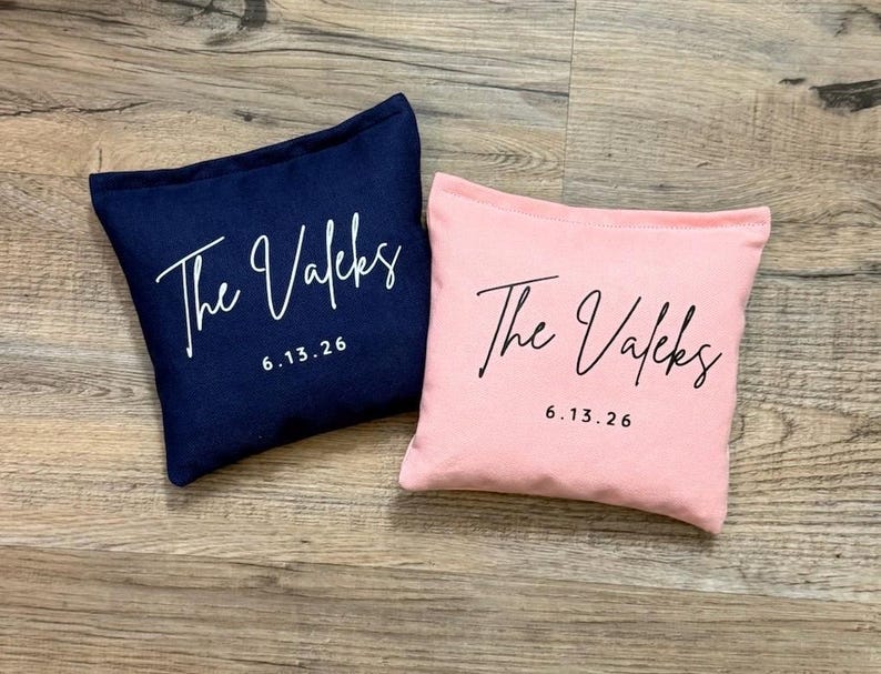 Puede incluir: Dos cojines decorativos cuadrados. Uno es azul marino, el otro es rosa claro. Ambos cojines tienen el texto "The Valeks" en escritura blanca y la fecha "6.13.26" en una fuente sencilla.