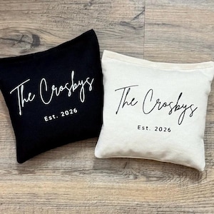 Puede incluir: Dos cojines cuadrados, uno negro y otro blanquecino, con el texto "The Crosbys" en cursiva y "Est. 2026" impreso debajo. Los cojines est&aacute;n colocados sobre una superficie de madera.