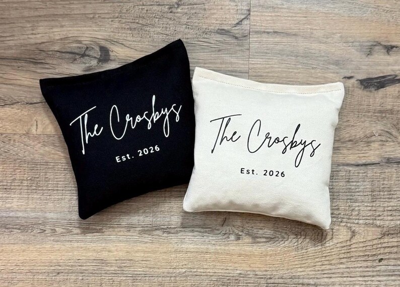 Puede incluir: Dos cojines cuadrados, uno negro y otro blanquecino, con el texto "The Crosbys" en cursiva y "Est. 2026" impreso debajo. Los cojines est&aacute;n colocados sobre una superficie de madera.