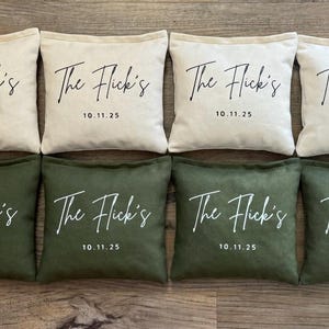 Puede incluir: Ocho sacos de frijoles cuadrados, cuatro en crema y cuatro en verde oliva. Cada bolsa tiene el texto "The Flick's" y la fecha "10.11.25" impresos en escritura blanca. Las bolsas est&aacute;n dispuestas en dos filas de cuatro.