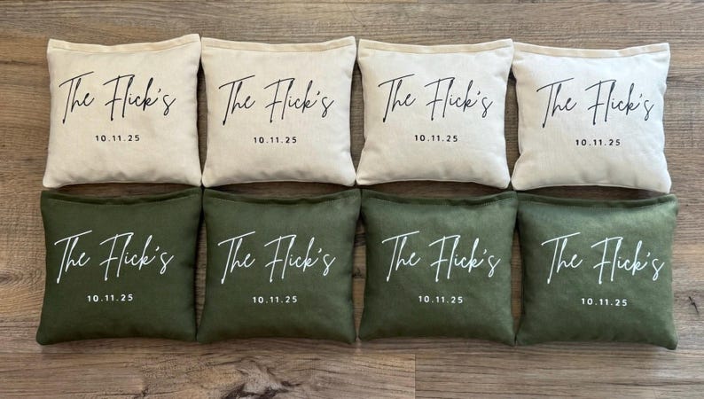 Puede incluir: Ocho sacos de frijoles cuadrados, cuatro en crema y cuatro en verde oliva. Cada bolsa tiene el texto "The Flick's" y la fecha "10.11.25" impresos en escritura blanca. Las bolsas est&aacute;n dispuestas en dos filas de cuatro.