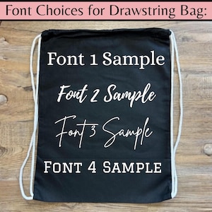 May include: A black drawstring bag with white text displaying font samples. The text includes "Font 1 Sample," "Font 2 Sample," "Font 3 Sample," and "FONT 4 SAMPLE." The bag has white drawstrings and is set against a wooden background.