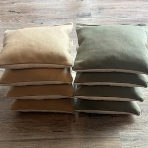 May include: A stack of eight bean bags, four are beige and four are olive green. The bean bags are made of canvas and have a white trim.