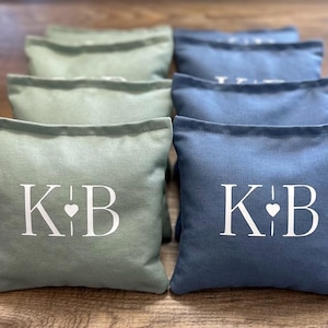 May include: A set of square bean bags in sage green and dusty blue fabric. Each bag features a white printed monogram with the letters K and B separated by a small heart symbol. The bags are arranged in two rows on a wooden surface.