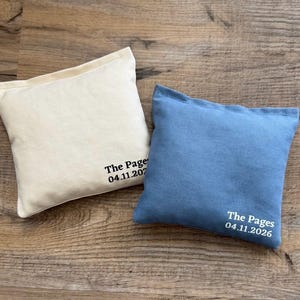 May include: Two square, small pillows. One is off-white, the other is blue. Both pillows have the text "The Pages" and a date printed in white. The pillows are on a wooden surface.