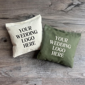 May include: Two square fabric pouches, one off-white with black text and one olive green with white text. Both pouches feature the printed words YOUR WEDDING LOGO HERE in a bold, sans-serif font, centered on the front of each fabric bag.