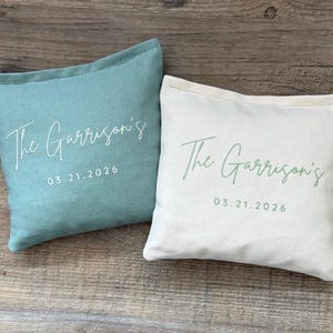 Puede incluir: Dos cojines cuadrados, uno turquesa y otro blanquecino, con el texto "The Garrison's" y la fecha "03.21.2026" impresos en letra cursiva. Los cojines est&aacute;n sobre una superficie de madera.
