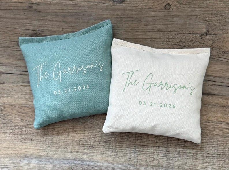 Puede incluir: Dos cojines cuadrados, uno turquesa y otro blanquecino, con el texto "The Garrison's" y la fecha "03.21.2026" impresos en letra cursiva. Los cojines est&aacute;n sobre una superficie de madera.