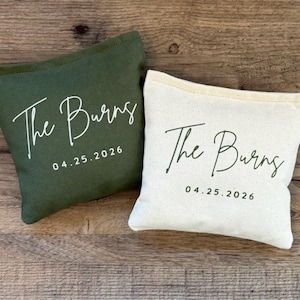 Puede incluir: Dos peque&ntilde;as bolsas rectangulares de lona. Una es verde oscuro, la otra es blanquecina. Ambas bolsas tienen el texto "The Burns" y la fecha "04.25.2026" impresos en cursiva. Las bolsas est&aacute;n sobre una superficie de madera.
