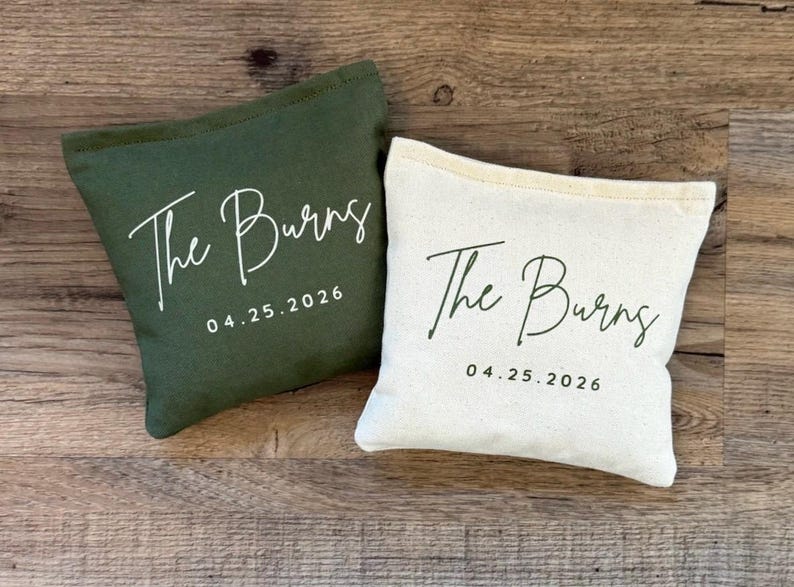 Puede incluir: Dos peque&ntilde;as bolsas rectangulares de lona. Una es verde oscuro, la otra es blanquecina. Ambas bolsas tienen el texto "The Burns" y la fecha "04.25.2026" impresos en cursiva. Las bolsas est&aacute;n sobre una superficie de madera.