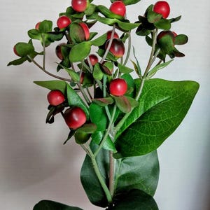 Może przedstawiać: Zbliżenie sztucznej kompozycji kwiatowej z żywymi czerwonymi jagodami i bujnymi zielonymi liśćmi. Jagody są okrągłe i błyszczące, kontrastując z ciemnozielonymi liśćmi. Łodygi są w stonowanej zieleni, tworząc naturalistyczny wygląd.
