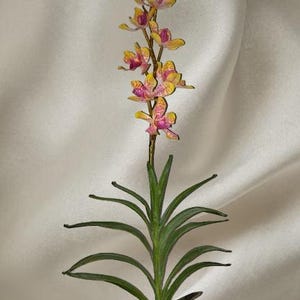 May include: A decorative artificial orchid stem featuring a vertical arrangement of small, vibrant yellow and magenta patterned flowers. The plant has a slender green stalk and several long, pointed, dark green leaves extending from the base.