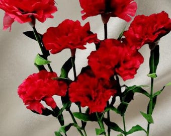 May include: A bouquet of vibrant red carnations with green stems and leaves. The flowers are in full bloom, showcasing their ruffled petals. The background is a soft, neutral fabric.