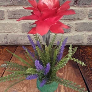 Pode incluir: Um vibrante arranjo floral artificial com uma grande flor vermelha com um centro rosa claro, rodeada por folhagem verde e roxa. O arranjo está num pequeno vaso verde, criando uma exibição colorida.