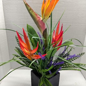 May include: A vibrant floral arrangement featuring a bird of paradise flower with orange and yellow petals, complemented by red flowers, green foliage, and purple accents. The arrangement is set in a black square vase.