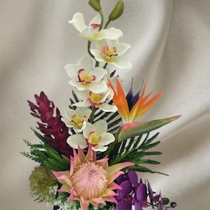 Puede incluir: Un vibrante arreglo floral con orquídeas blancas, una flor de ave del paraíso y una protea rosa. El arreglo incluye follaje verde y está colocado en una base cuadrada negra.
