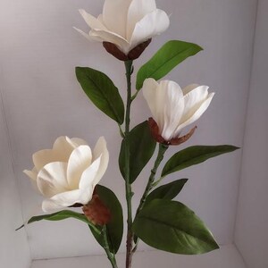 Puede incluir: Un tallo de flor de magnolia artificial blanco con tres flores y hojas verdes.