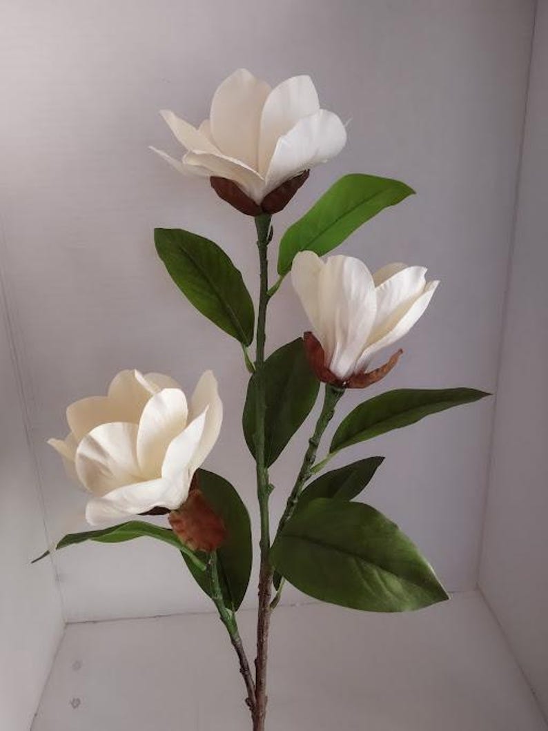 Puede incluir: Un tallo de flor de magnolia artificial blanco con tres flores y hojas verdes.